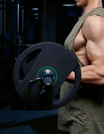Side view of incognito bodybuilder training biceps with barbell. Crop of muscular sportsman with perfect body posing in gym near mirror and dumbbells in dark atmosphere. Concept of bodybuilding.