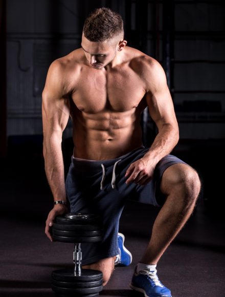 Portrait of young attractive caucasian muscular man working out in gym, leaning on dumbbell, showing chest, shoulder, abs muscles